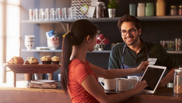 Male barista with beard and glasses standing behind counter while brunette woman with ponytail uses credit card to pay at Bank of Utah merchant services card reader.