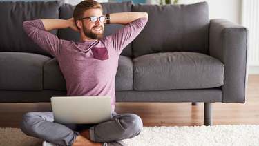 Smiling, relaxed man sits in front of couch, with laptop open on his lap