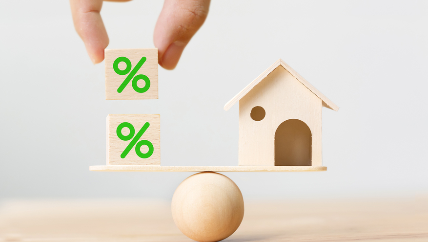 A toy wooden house sits on one side of a balance while a hand drops wooden blocks with percent signs on the other side