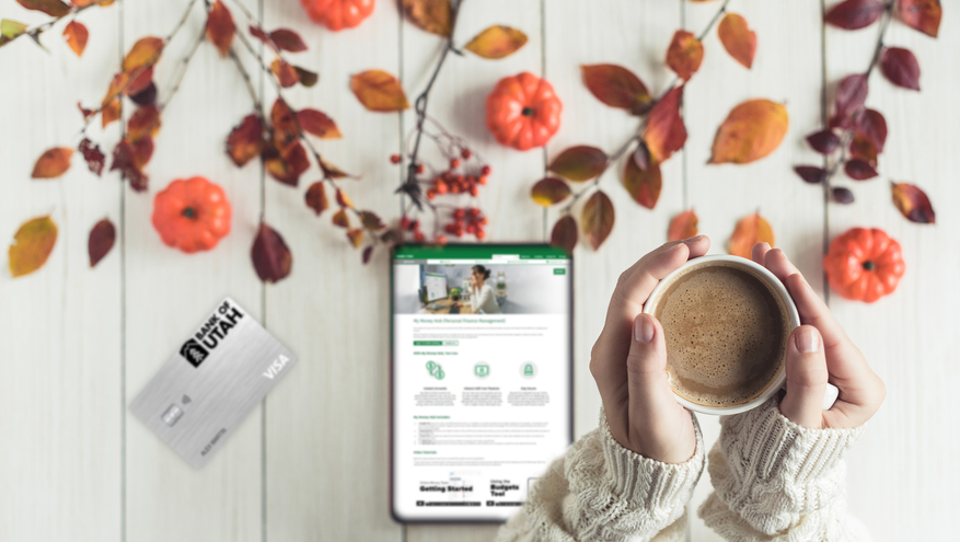 A pair of hands hold a hot chocolate mug over a fall-themed table with an iPad and Bank of Utah credit card on it