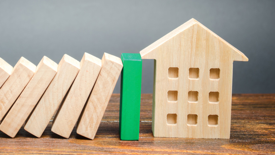 An image of wooden blocks toppling toward a wooden house, with a green block stopping them from falling