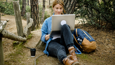 A college-age female works on her laptop in a wooded setting