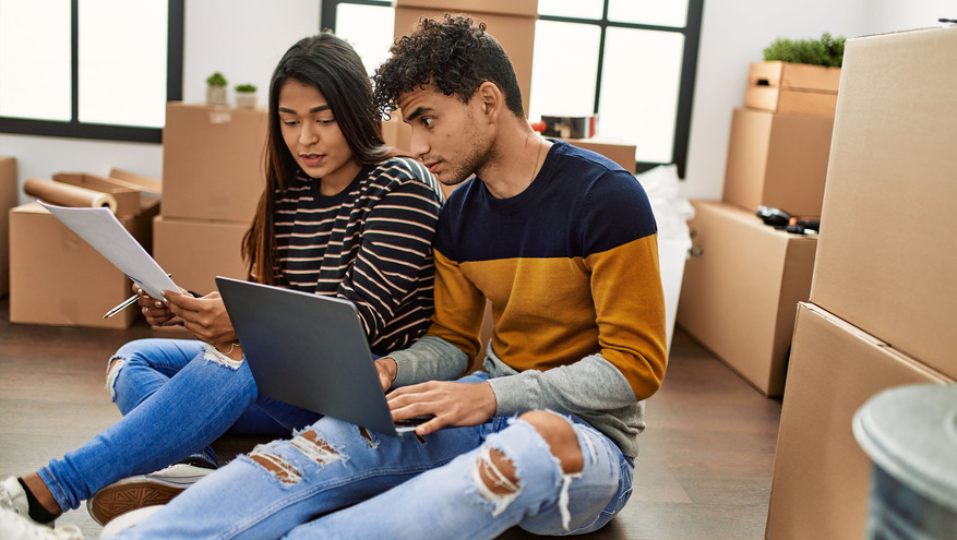 A young, diverse couple review paperwork while sitting among moving boxes