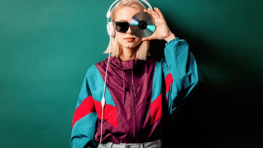 A women wearing a 1990s era wind jacket and acid wash jeans holds up a music CD over her eye