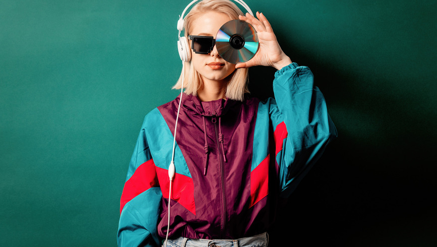 A women wearing a 1990s era wind jacket and acid wash jeans holds up a music CD over her eye