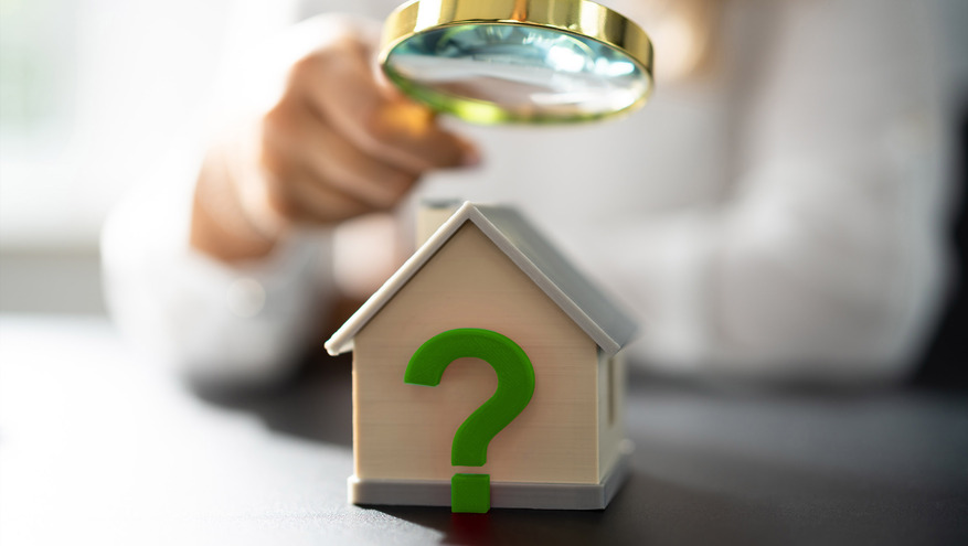 A blurred person holds a magnifying glass over a miniature wooden house with a question mark on it
