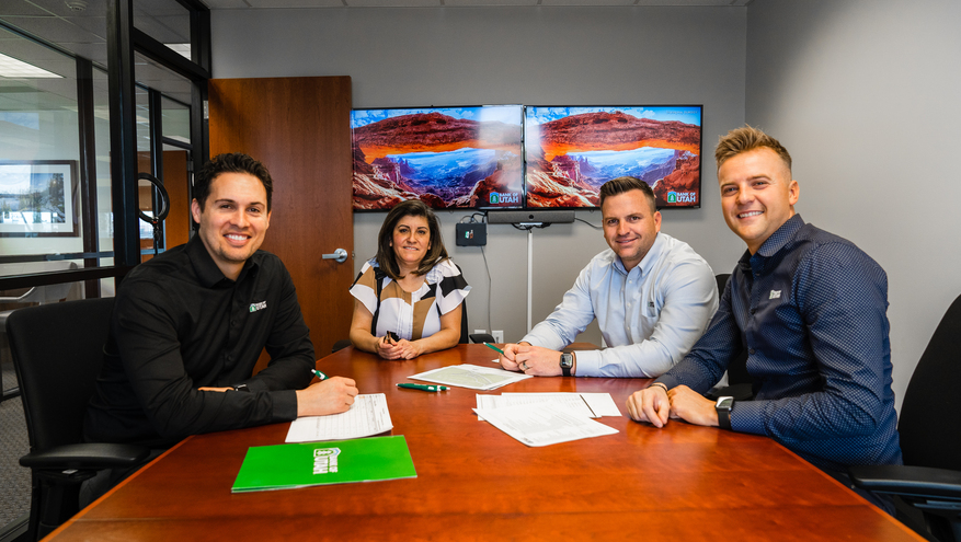 Four Bank of Utah team members sit around a conference table, smiling toward the camera. Documents and branded folders are on the table, and two monitors on the wall behind them display scenic Utah landscapes with the Bank of Utah logo.