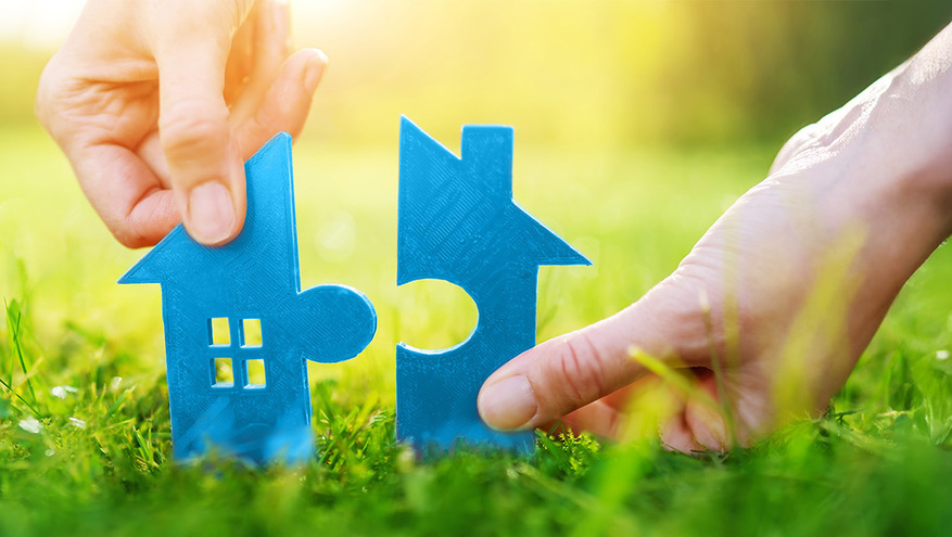 Two hands fitting together blue house-shaped puzzle pieces on green grass, symbolizing the perfect mortgage or home loan solution from Bank of Utah.