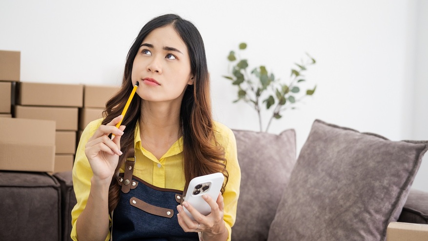Thoughtful small business owner sitting on a couch with shipping boxes in the background, holding a pencil and smartphone, appearing to consider a decision.