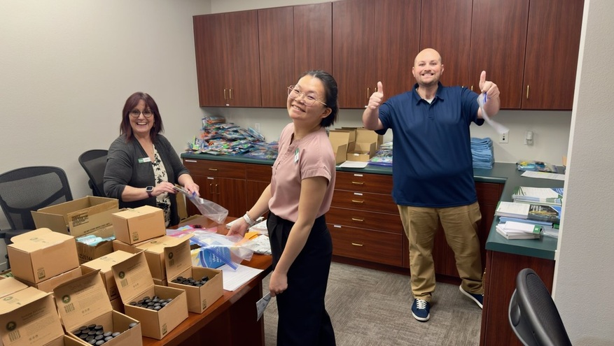 Three Bank of Utah team members smile and give thumbs up while assembling hygiene kits in an office. The table is filled with open boxes of supplies, and completed kits are stacked in the background