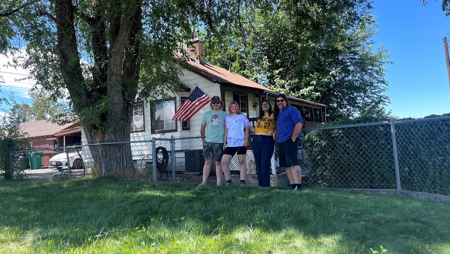 A family of four stands together smiling in front of a small house with an American flag on the porch, shaded by large trees and a chain-link fence in the foreground.