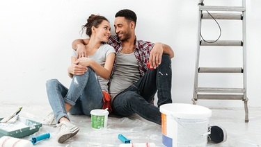 A smiling couple sits on a drop-cloth-covered floor among paint buckets and a ladder while taking a break from painting a white room.