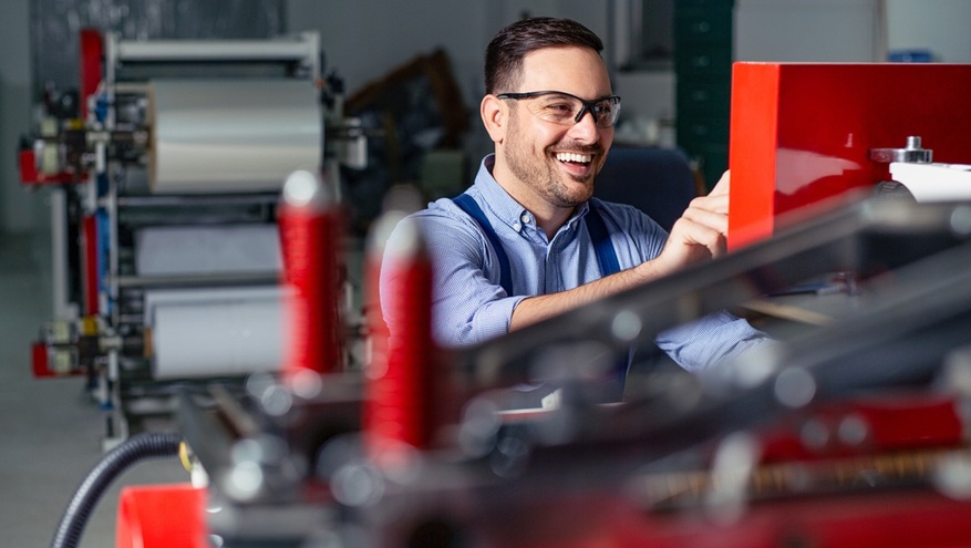 A smiling male factory worker wearing safety glasses and overalls operates a large piece of manufacturing machinery. In the background, industrial equipment and large rolls of material are visible in a brightly lit facility.