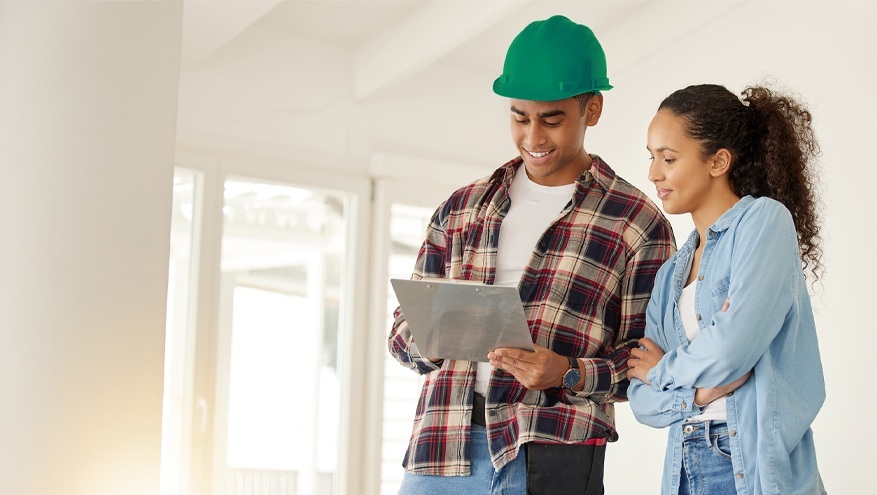 A construction contractor in a green hard hat shows a clipboard to a woman inside a brightly lit building that's under construction