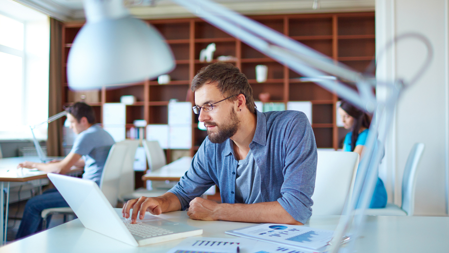 Thin man wearing glasses working on laptop in an open office setting with coworkers in background.