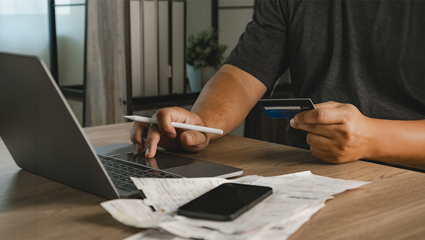 Close-up of a person at a wooden desk holding a blue business credit card in one hand and using a laptop touchpad with a stylus in the other, next to a stack of paper receipts and a mobile phone.