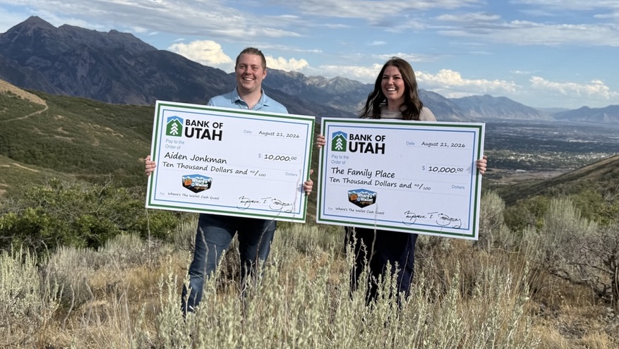 Where’s the Wallet winner Aiden Jonkman and a representative standing outdoors on a mountain trail, holding two $10,000 Bank of Utah novelty checks made out to Aiden and The Family Place.