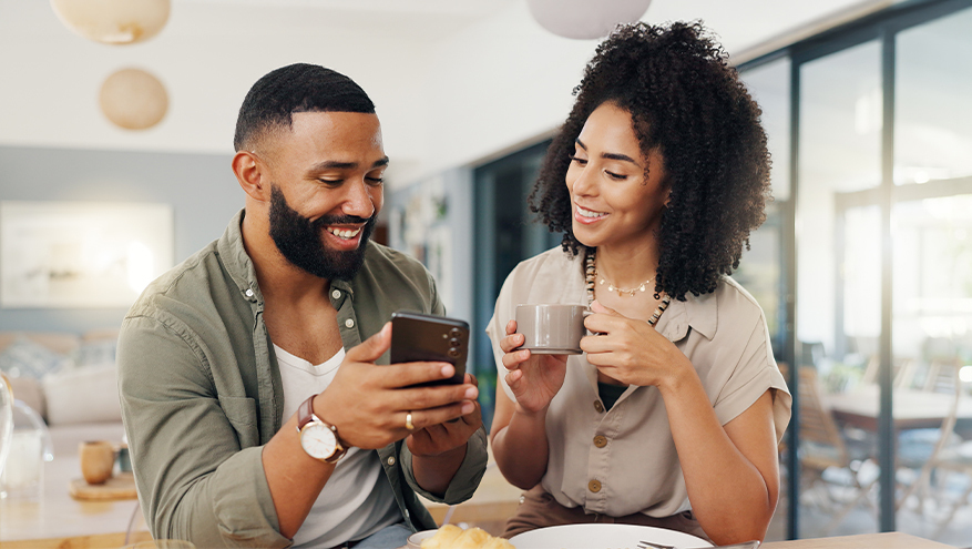 A smiling couple sitting together as the man shows something on his smartphone while the woman holds a coffee mug and looks at the screen.