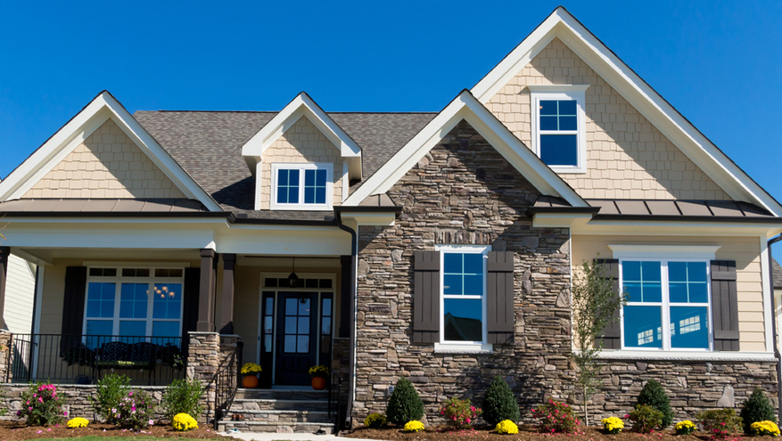 Front view of new house made of rock and cream colored siding, with six front facing windows. Green front yard with new flowers, plants and trees on blue sky background.