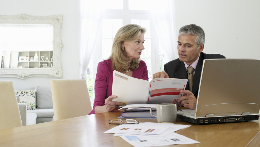 Elderly woman going over finances with male financial advisor. Sitting at dining room table with laptop and financial papers.