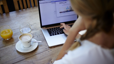 Woman working on laptop at kitchen table with cup of coffee and glass of orange juice.