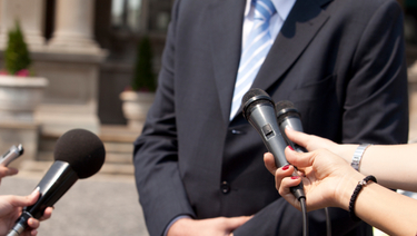 Four arms outstretched holding microphones to man in suit, that is standing in front of mansion.