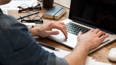 Man working at desk on laptop. Eyeglasses, mug, book and papers on desk.