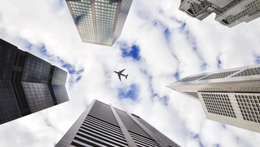 Point of view is looking up amidst tall city buildings with white clouds over blue sky and airplane flying overhead.