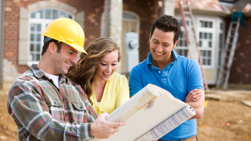Young couple looking at blueprints with construction worker in front of new house being built.