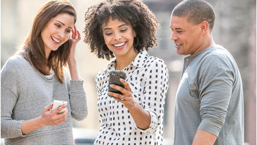 Three young adults smiling and looking at smartphone being help by woman standing in the middle.