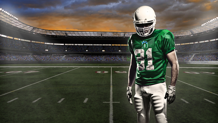 Football player in full pads wearing a green uniform with Bank of Utah logo, standing alone on football field, surrounded by bleachers full of fans.