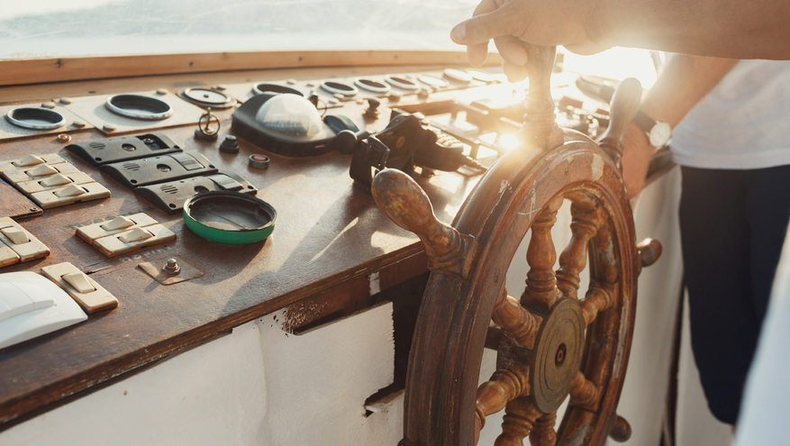 Hand on ship steering wheel or helm, with navigation gauges and tools on dashboard.