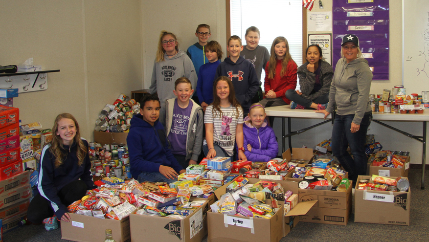 Group on children with teacher surrounded by boxes of non-perishable foods for the 2018 Kick Childhood Hunger Food Drive.