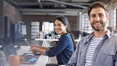 Young man and woman working in an open office setting with two open laptops.