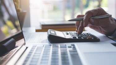 Close up of hand holding a pen and using a calculator on a desk that is sitting next to an open laptop.