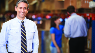 Man dressed in business clothes standing in warehouse with coworkers working in the background.