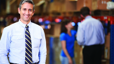 Man dressed in business clothes standing in warehouse with coworkers working in the background.