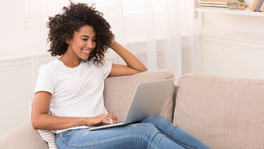 Happy young woman with dark hair sitting on couch working on a laptop.