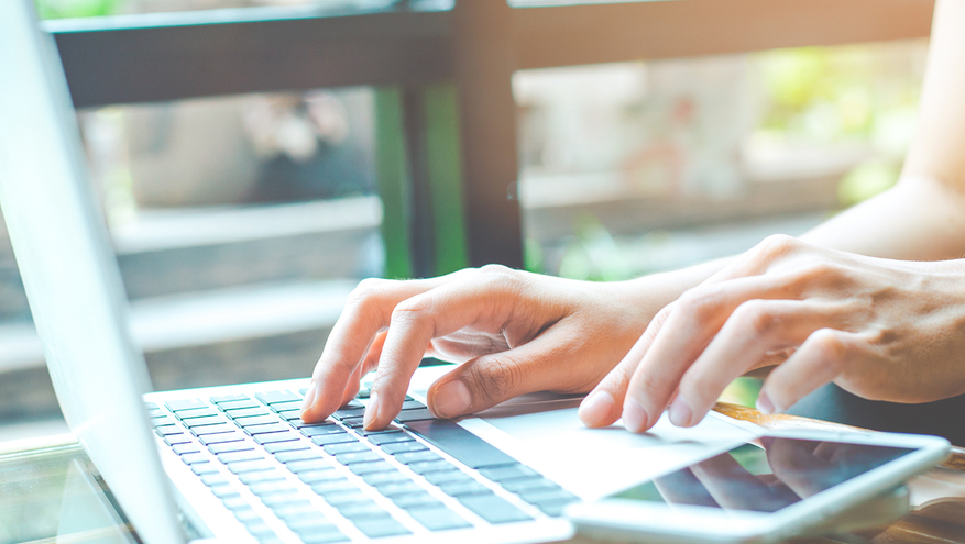 Close up of woman's hands typing on laptop on desk with smart phone next to laptop.