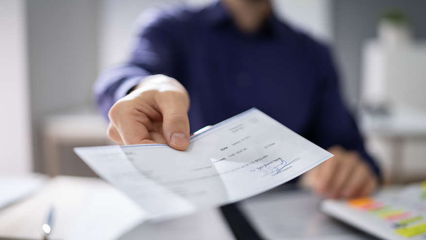 Close up of printed check being held out by a man's had over office desk.