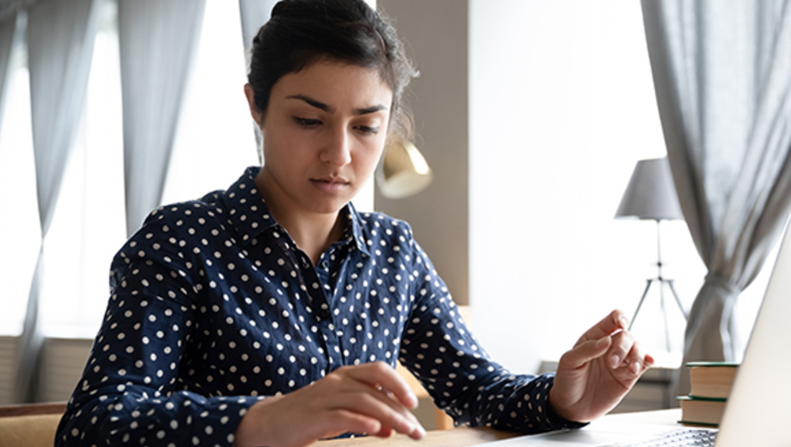 Woman in polka dot dress typing on calculator at desk next to a laptop.