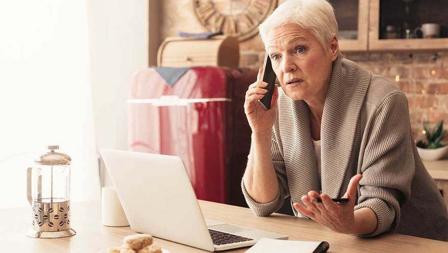 Senior woman having a serious telephone conversation in her kitchen, with her laptop open on a table