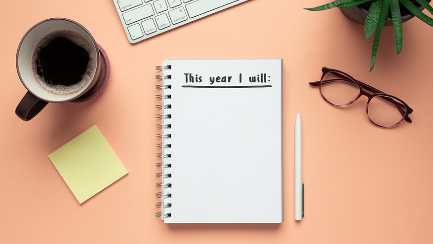 A notepad with "This year I will:" written on it sits on a desktop with a cup of coffee, keyboard, pen, post-its and a plant