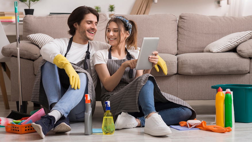 A young couple sits on the floor in front of their couch, looking at a tablet. They are wearing cleaning gloves and have cleaning products on the floor around them.